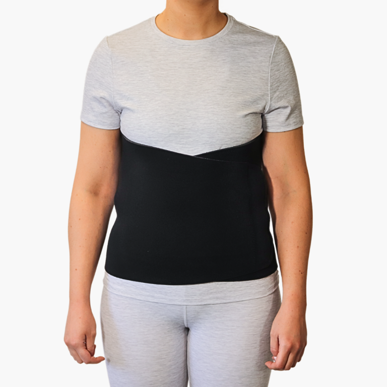 A woman wearing a wide black TheraCore® Abdominal Binder over a grey t-shirt for post-surgery core support and compression.